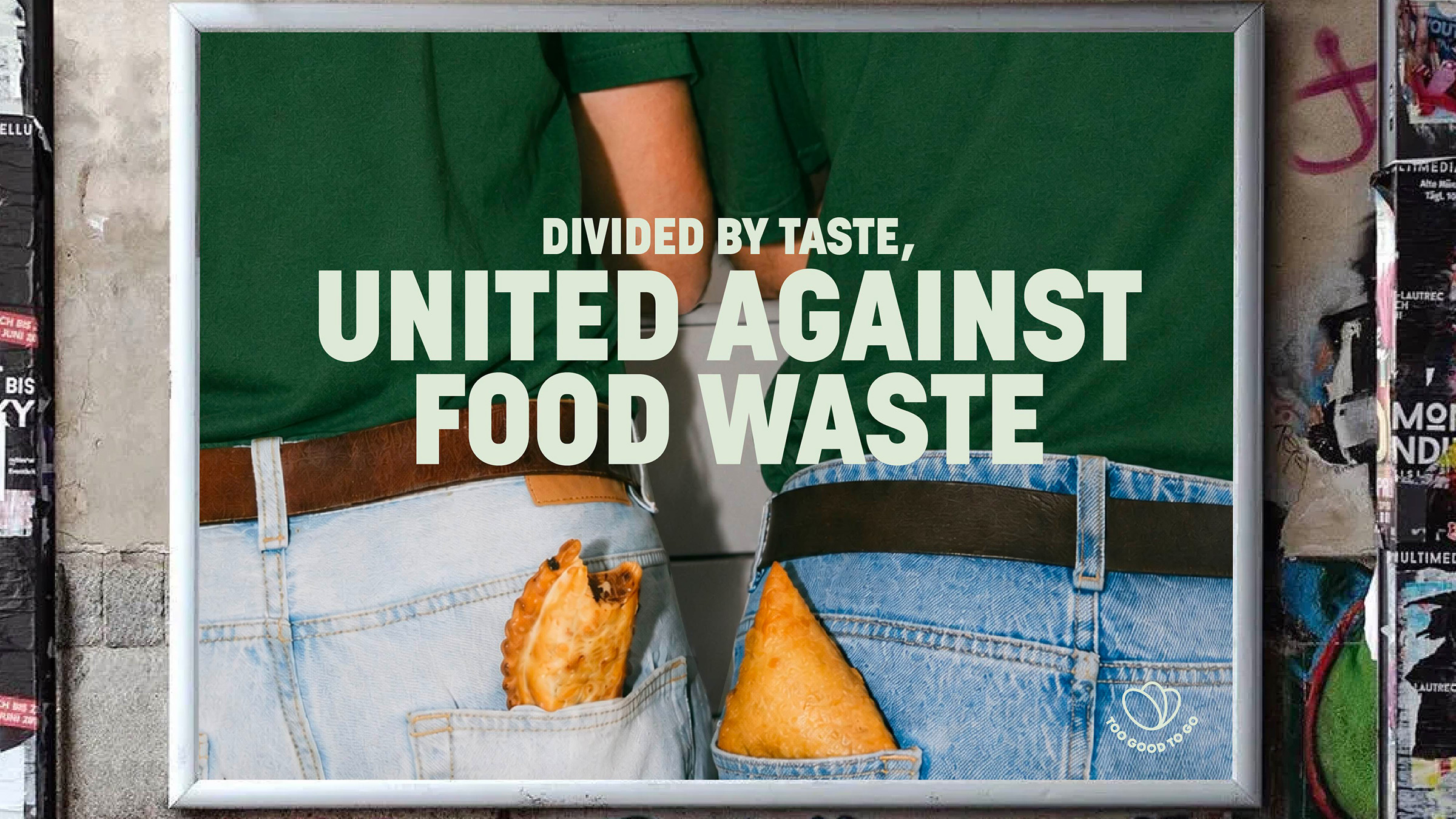 A framed poster on a street wall reading ‘Divided by taste, United against food waste’, over a photo of two people with pastries in their back pockets