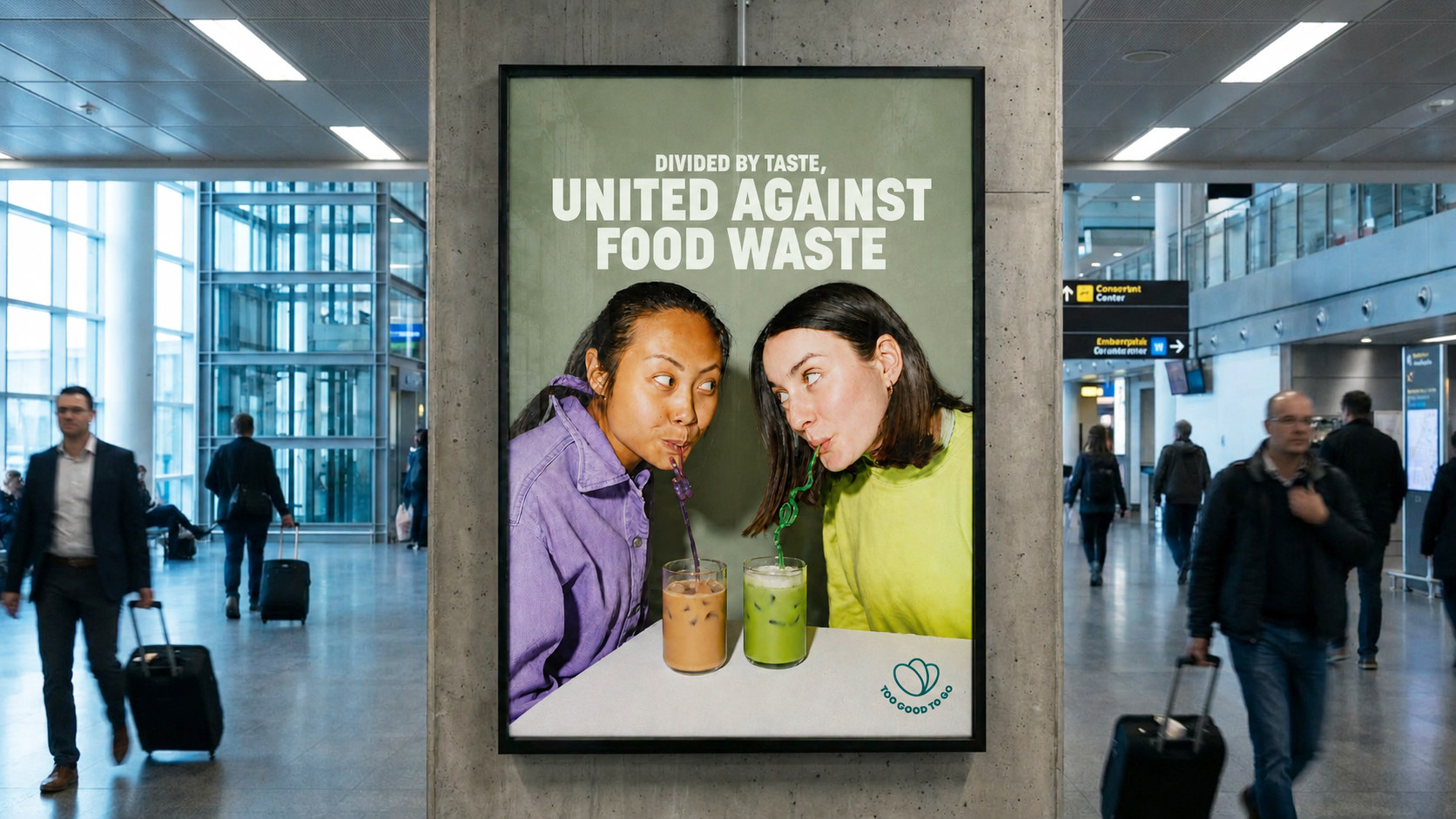 An airport poster of two women drinking iced coffee and matcha through straws, reading ‘Divided by taste, United against food waste’